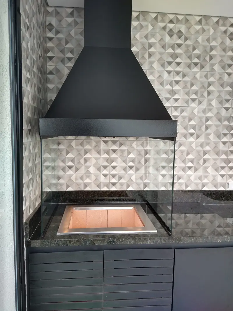 Churrasqueira gourmet com coifa preta, braseiro e vidro lateral, em área com revestimento geométrico