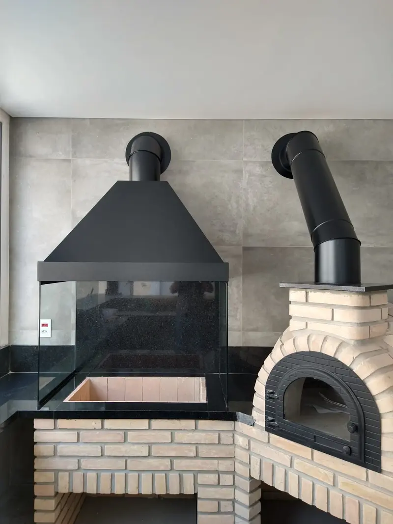 Churrasqueira com coifa preta e vidro, ao lado de forno de pizza de tijolinho