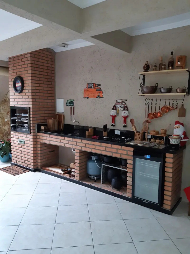 Área gourmet com churrasqueira de tijolinho, bancada de granito preto e geladeira pequena